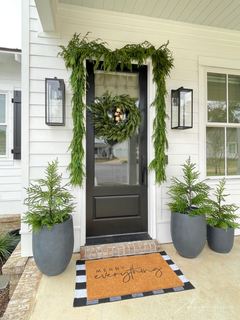 christmas holiday front door inspo from a white cottage with garland over the door and small trees in pots by door.  Norfolk pine trees and garland 