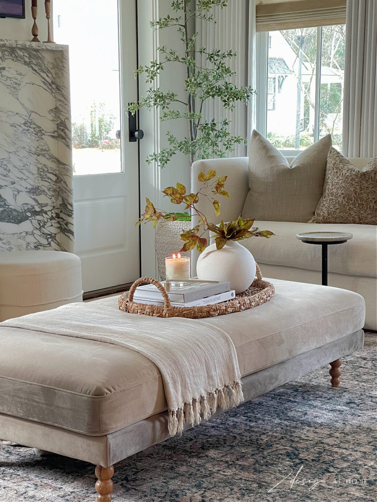 upholstered ottoman styled with basket trays, books, candle and white bubble vase with fall winter spring foliage, and a linen throw 